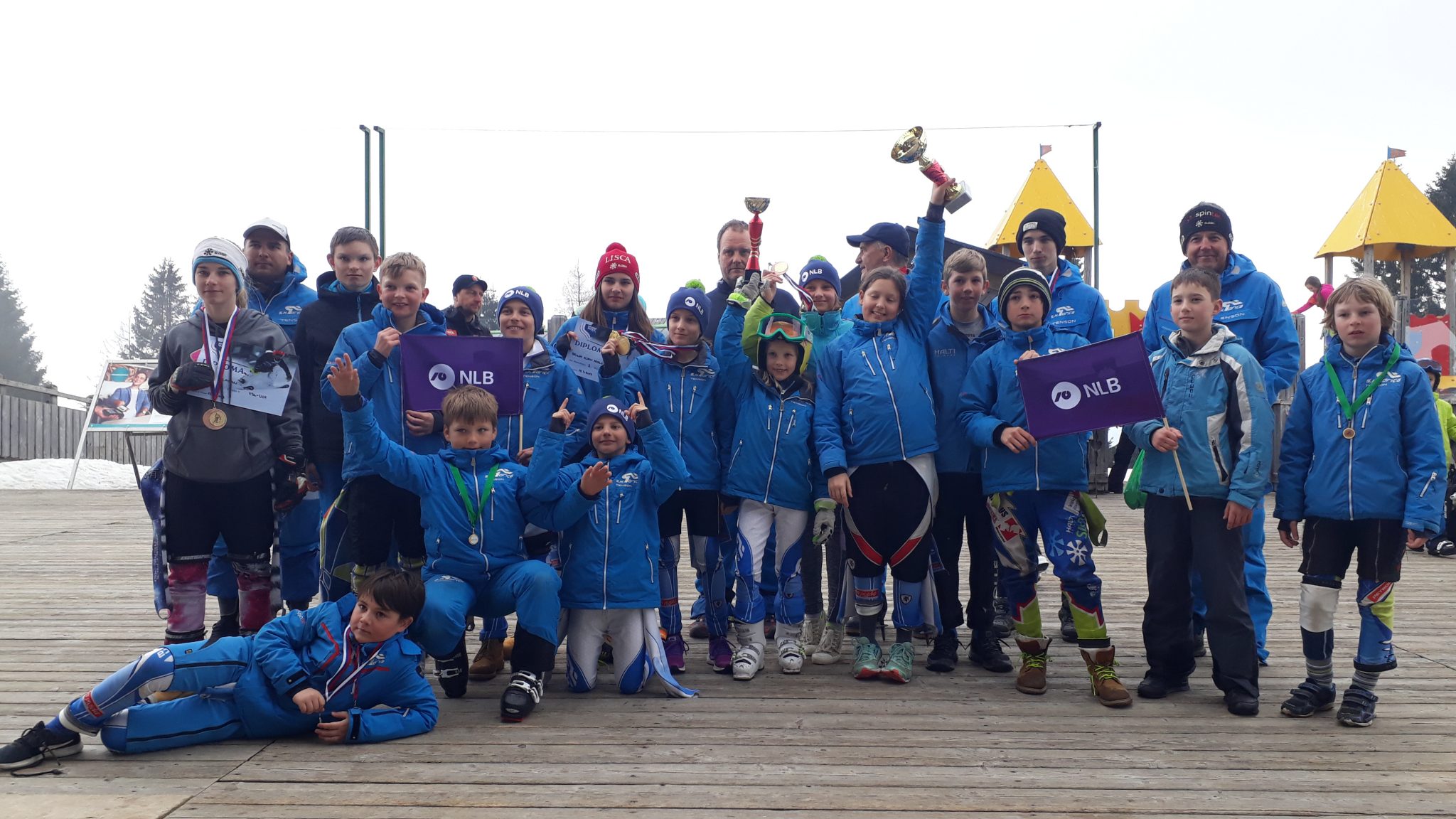 Skupina smučarjev različnih starosti, ki držijo nagrade, priznanja in zastave NLB